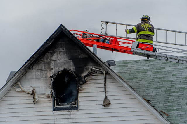 Damage caused by fire visible on the side of a house with a firefighter on a ladder.